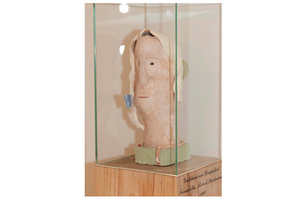 Skulptur eines stilisierten Kopfes mit Tuch in einer Glasvitrine auf einem Holzsockel